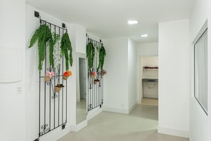 Un pasillo con paredes blancas, una zona de cocina al fondo y plantas decorativas en una reja de metal negro.