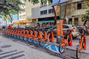 Una fila di biciclette arancioni parcheggiate su una strada acciottolata, un edificio con una tenda verde e un albero.