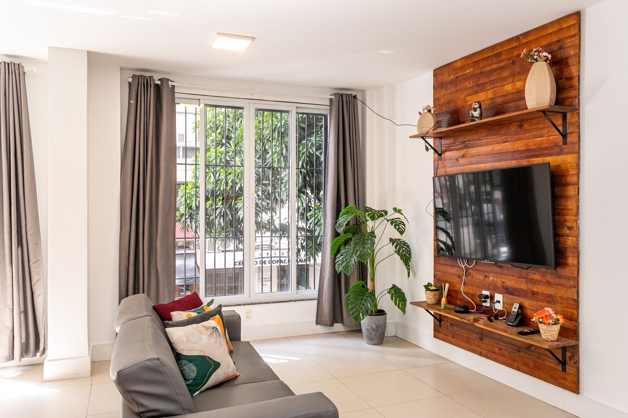 Un salon moderne avec un canapé gris, une grande plante en pot, un meuble TV en bois et une grande fenêtre avec des rideaux.