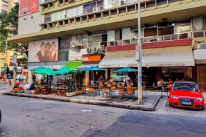 A street view with outdoor seating, a bakery, and a red car parked on the side. - Temporada Prime - Praia (Rio de Janeiro)