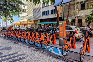 A row of orange bicycles parked along a cobblestone street, with a building featuring a green awning and a sign that reads "Nutmeg." - Temporada Prime - Praia (Rio de Janeiro)