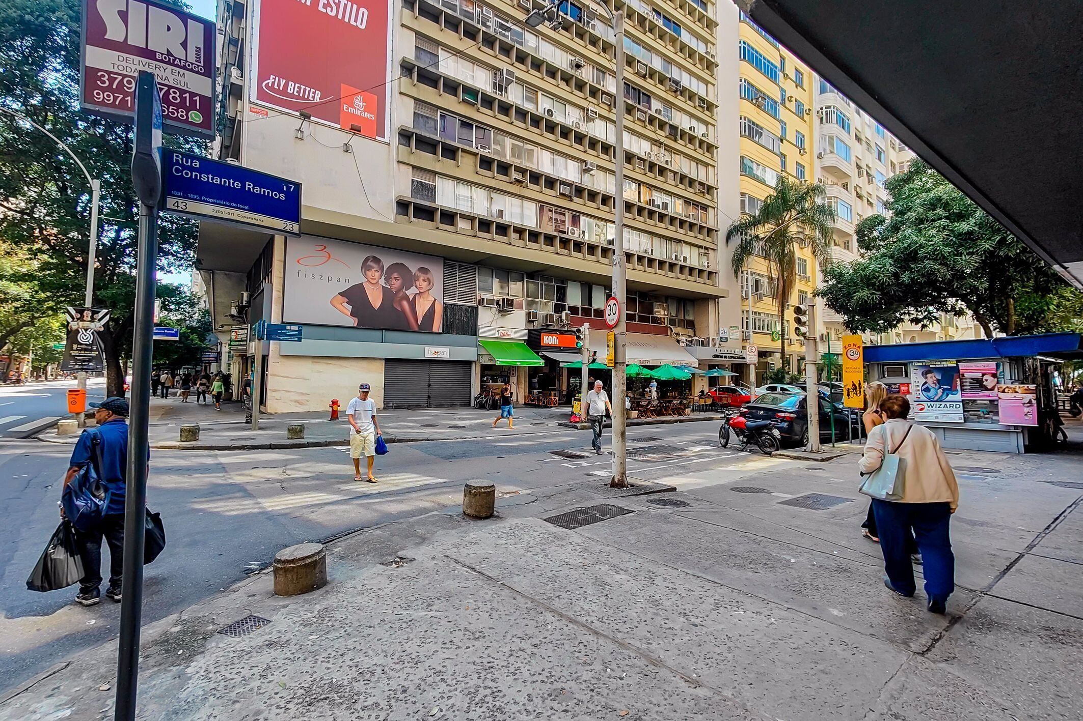 Une rue animée en milieu urbain, avec des piétons, un grand immeuble doté de nombreux balcons et divers commerces.