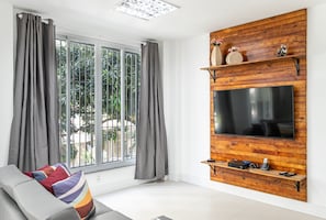 Un soggiorno moderno con una grande TV fissata a una parete in legno, tende grigie, un divano grigio con cuscini colorati e vista su piante.