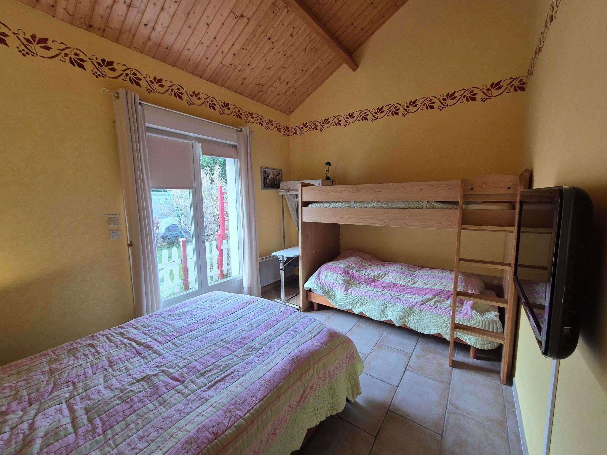 Una habitación con dos literas, techo de madera y ventana con vista a una valla roja y follaje exterior.