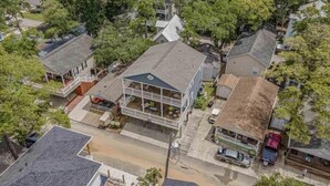 An aerial view of a residential area with houses, a prominent two-story house with a balcony, and a street with parked cars.