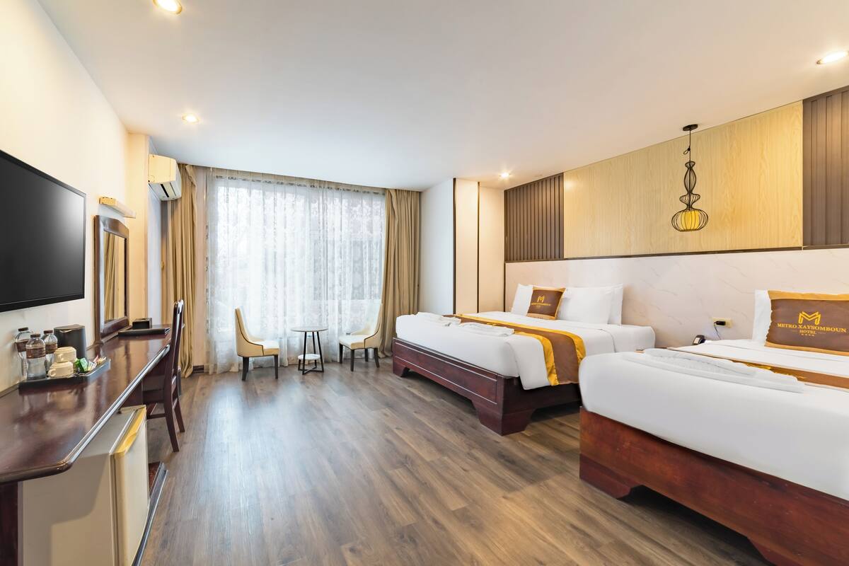 A hotel room with two beds, a desk, a TV, and a large window with curtains.