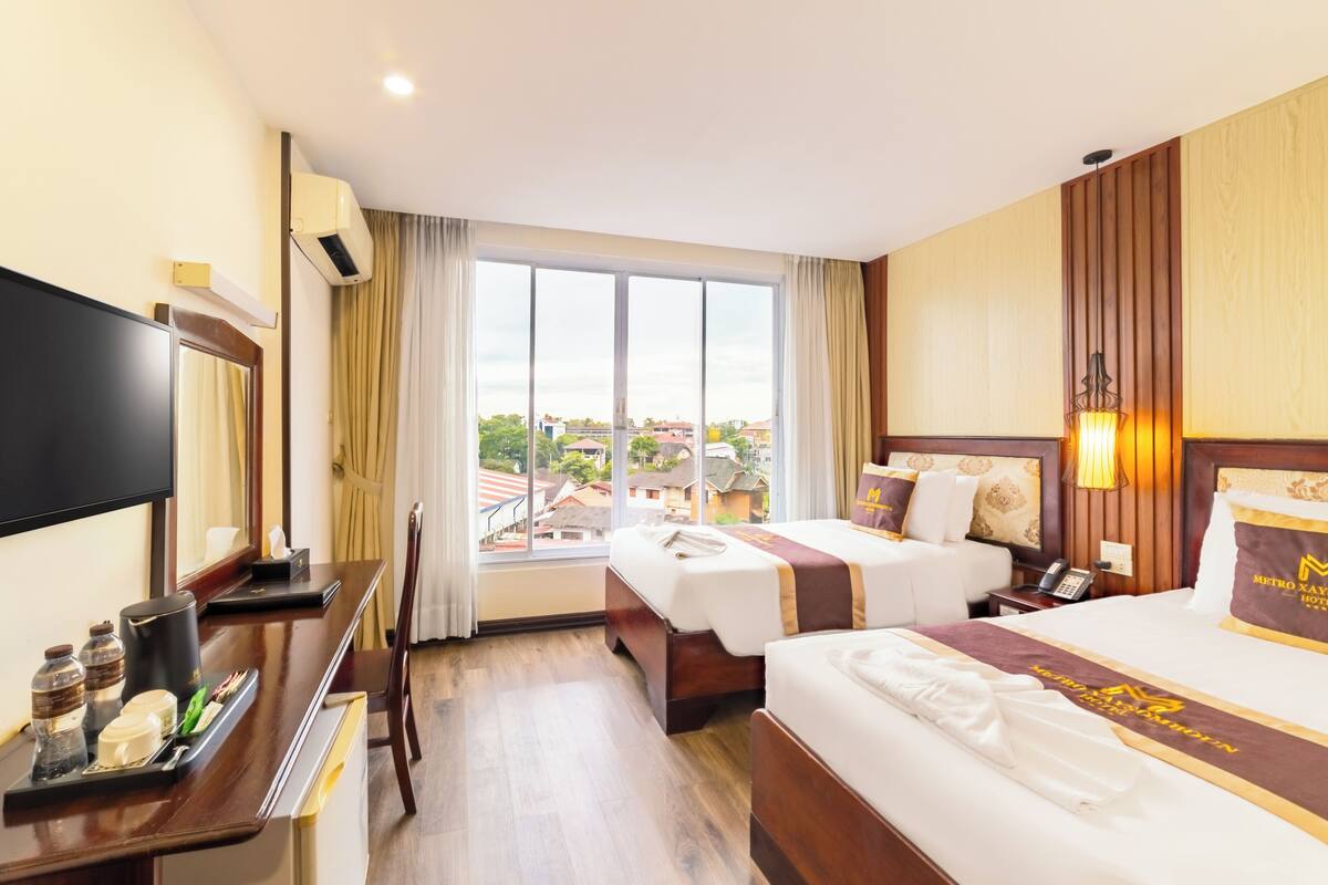 A hotel room with two beds, a desk with a flat-screen TV, a window with a view of buildings, and a wall-mounted air conditioner.