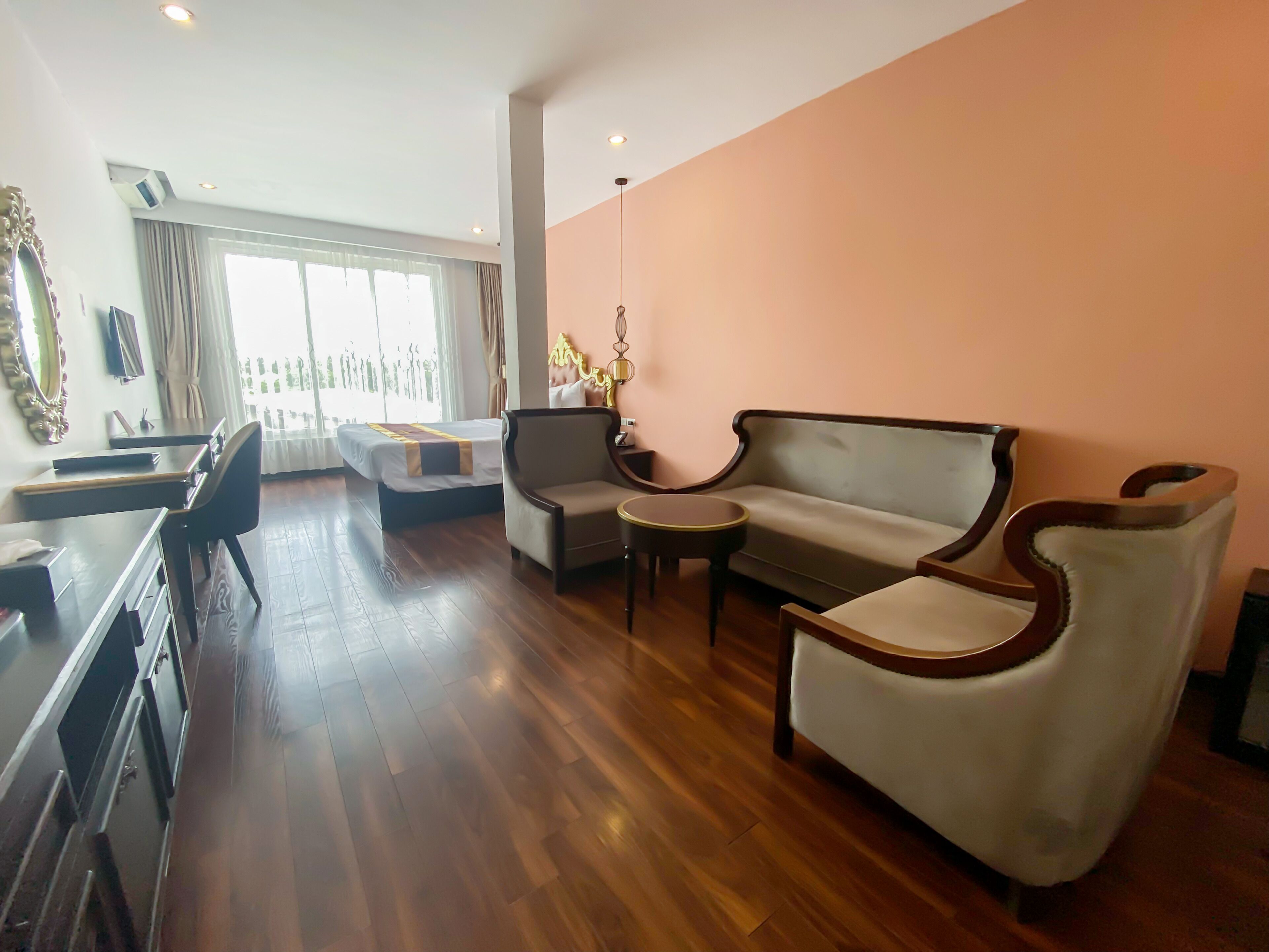 A hotel room with a wooden floor, a sofa, a chair, a small table, a desk, and a large window with curtains.