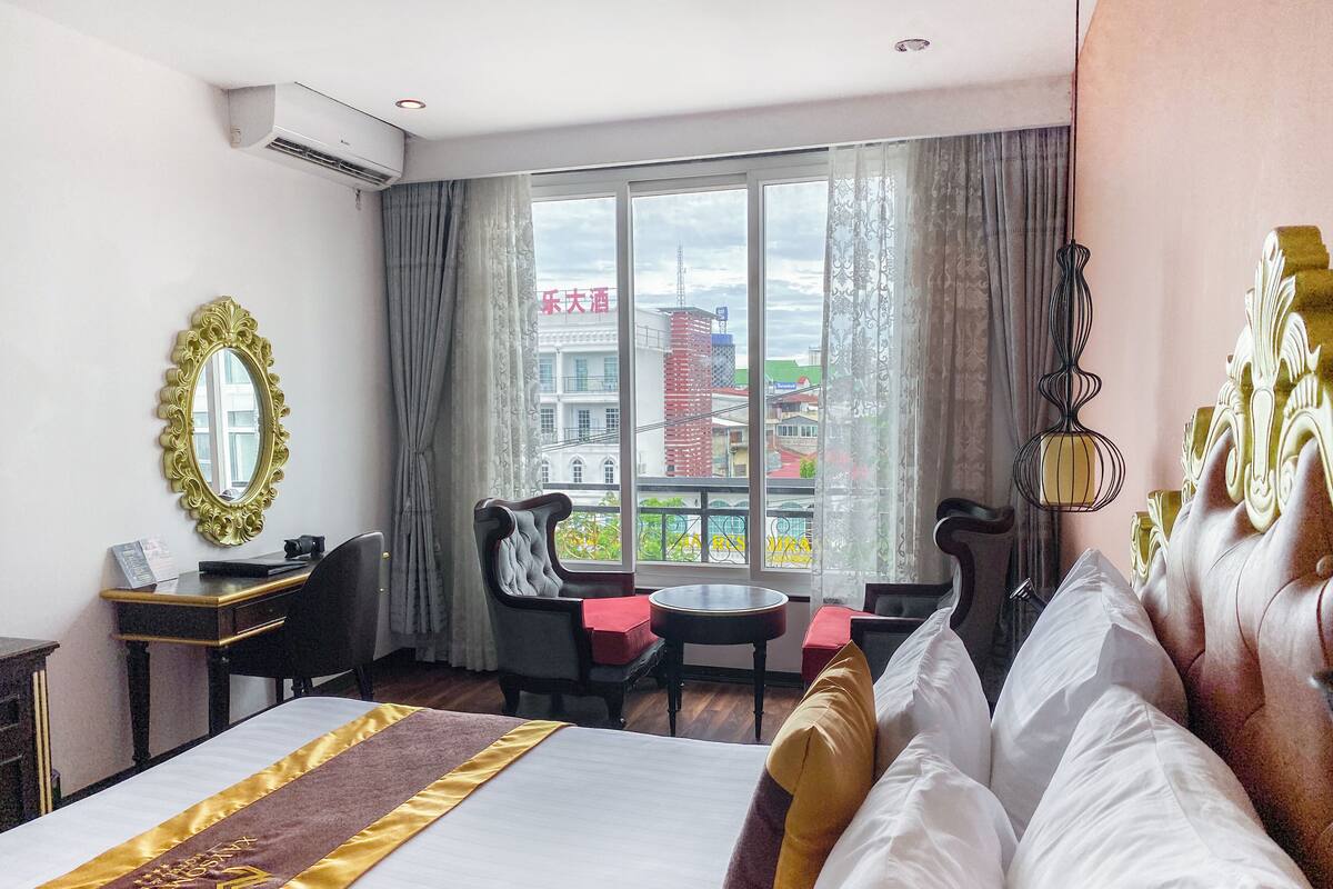 A hotel room with a large bed, a desk with a chair, a small table, and a view of the city through the window.