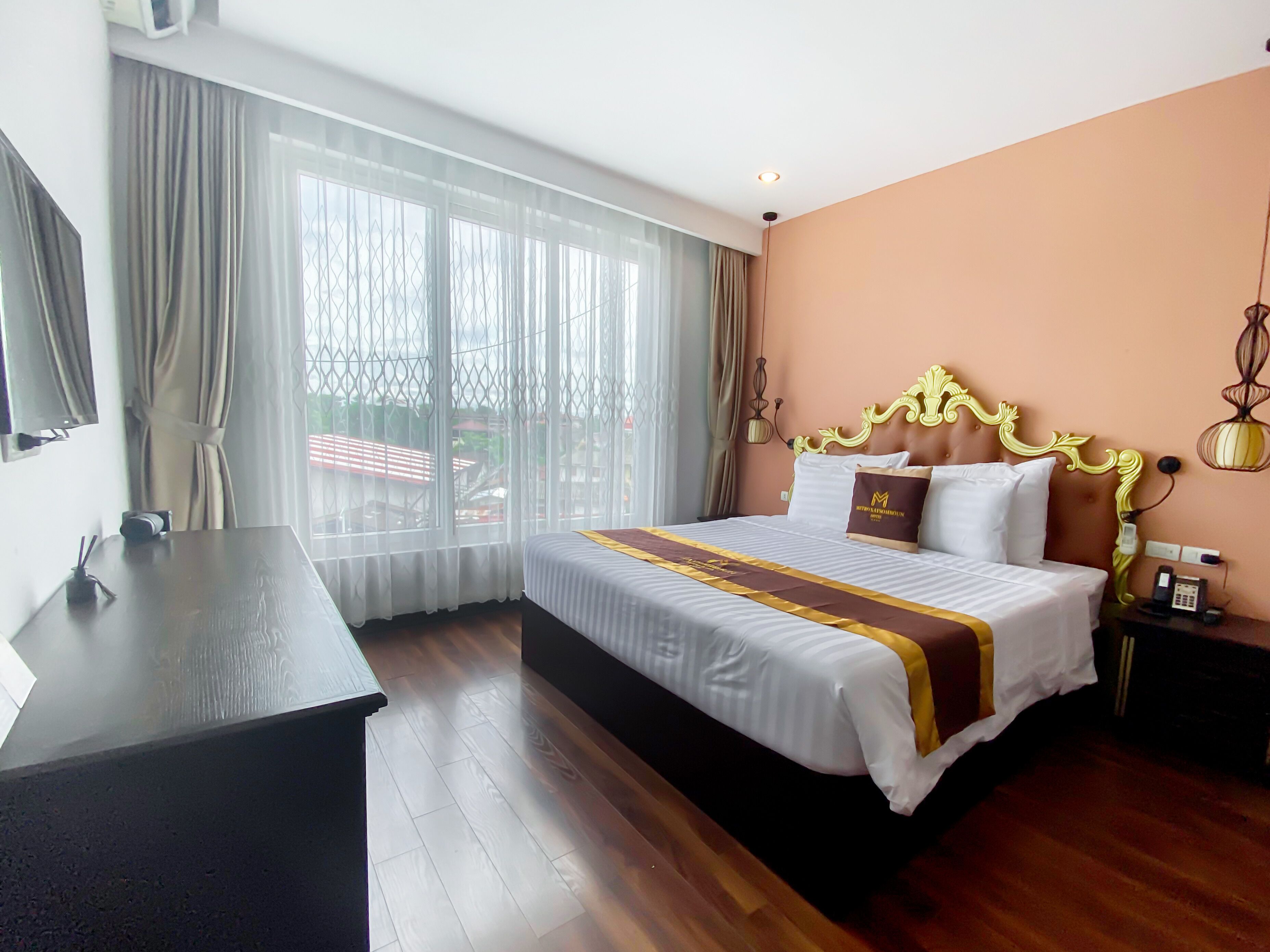 A hotel room with a large bed, a desk, and a view through sheer curtains.