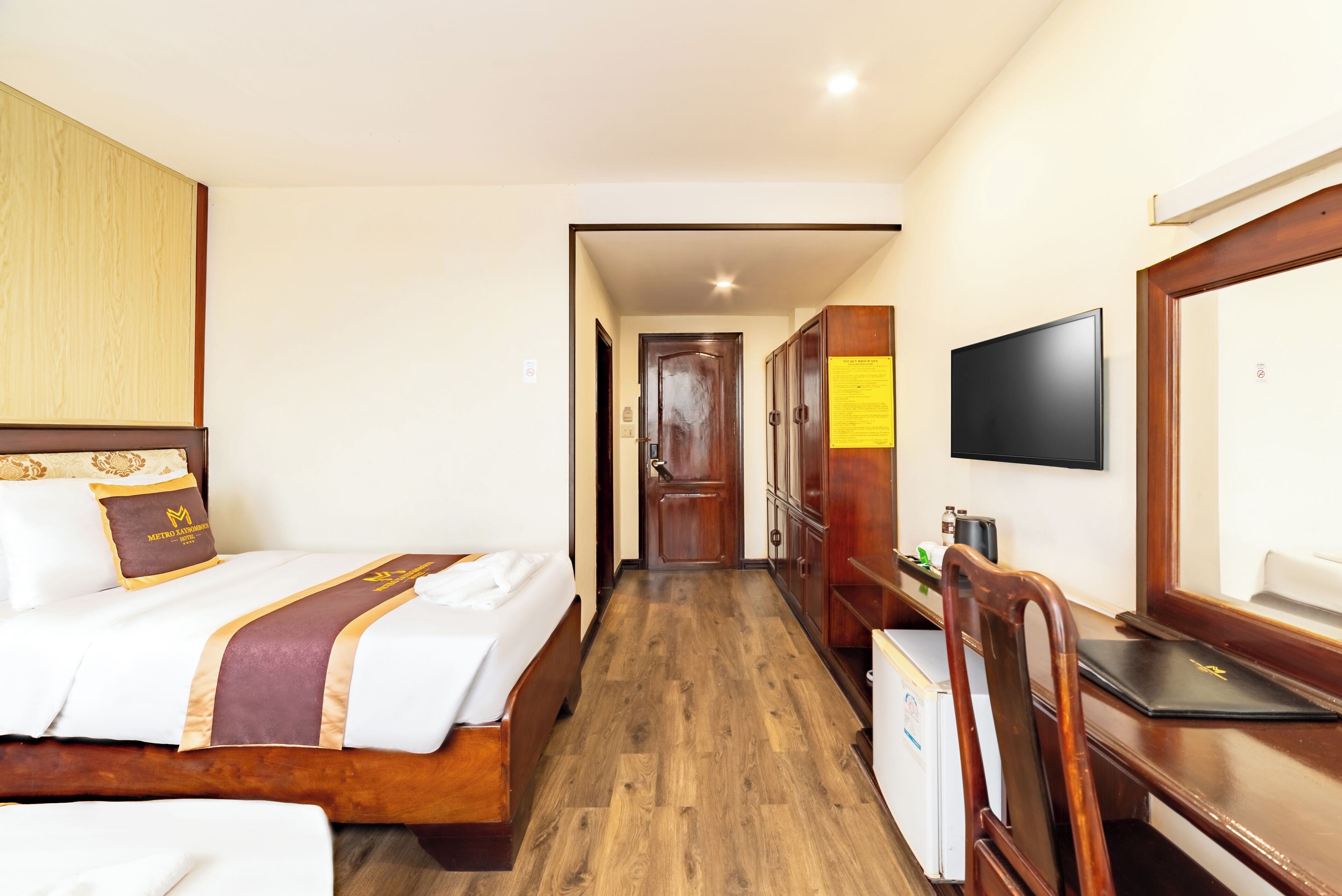 A hotel room with a wooden floor, a bed with a headboard, a desk with a chair, a television, and a wardrobe.