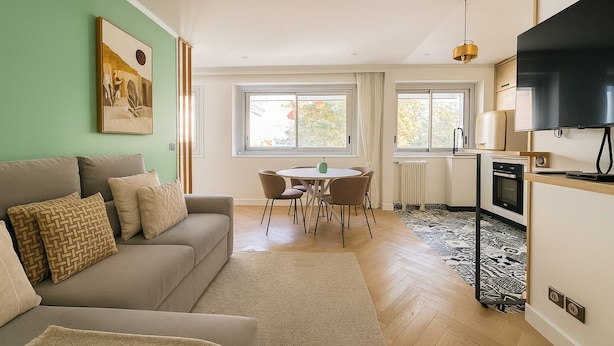 Una sala de estar moderna con sofá, una mesa de comedor con sillas, una zona de cocina con televisor y ventanas amplias.