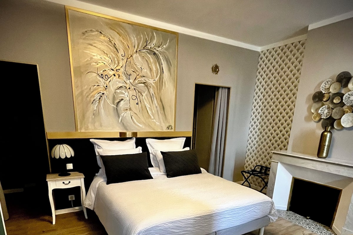 A modern hotel room with a large bed, a small side table, a chair, and a fireplace.