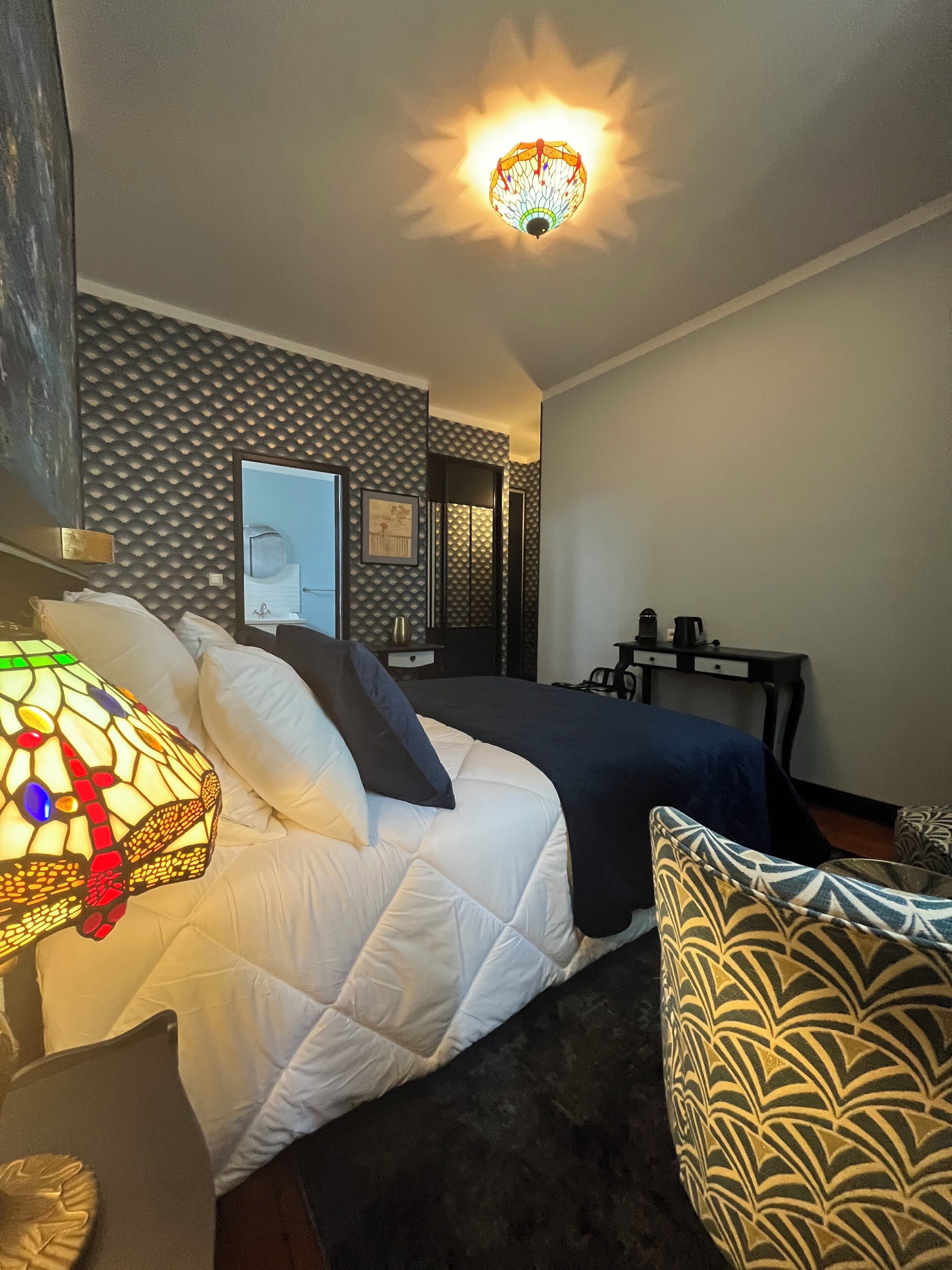 A hotel room with a bed, a nightstand with a lamp, a desk, and a chair.