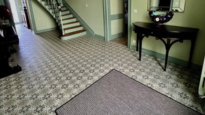 Una habitación amplia con piso estampado, una escalera, un piano, un espejo y una puerta que da a otra habitación.