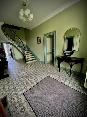 A spacious room with a patterned floor, a staircase, a piano, a mirror, and a door leading to another room. - Le Rococo (Couquèques)
