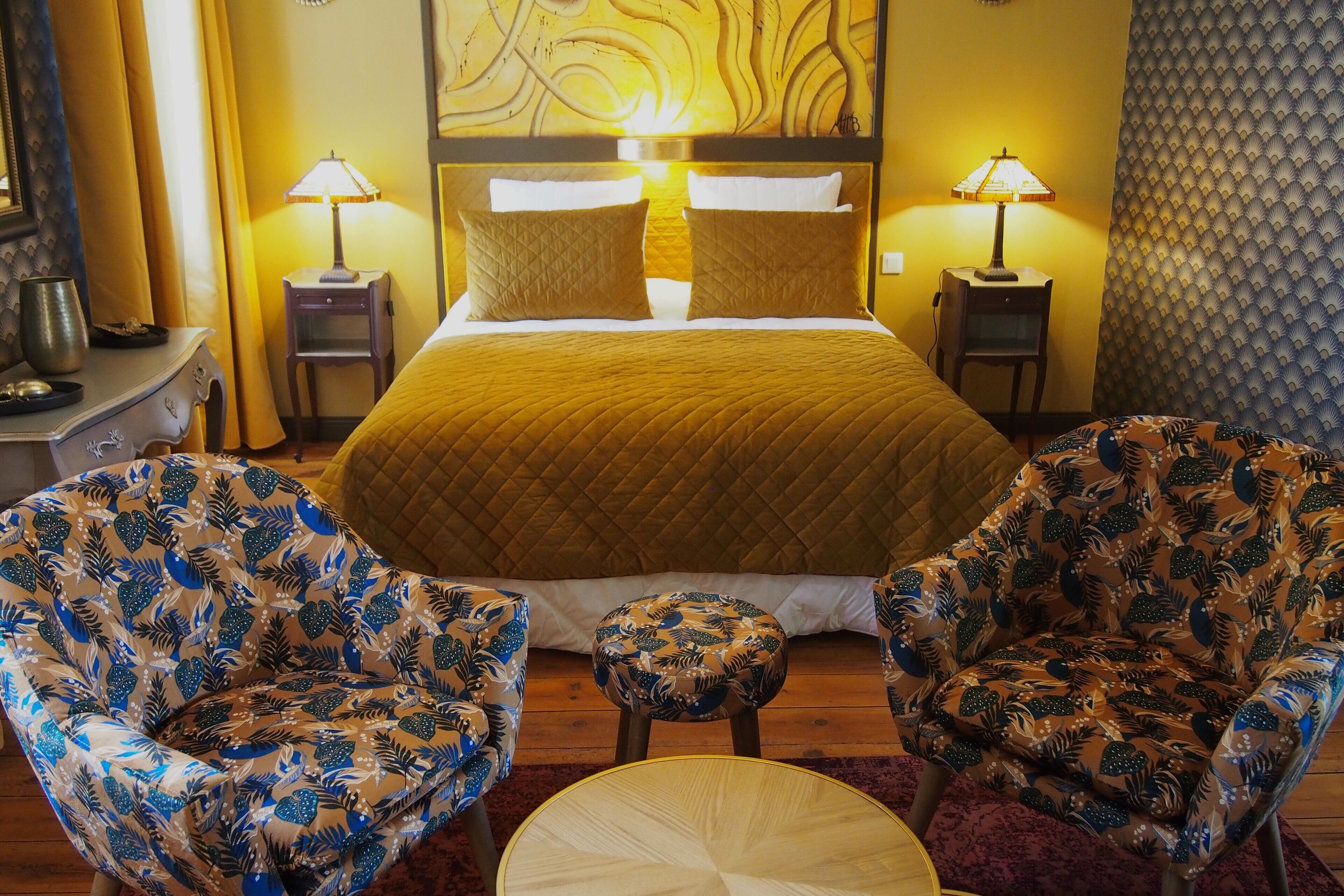 A hotel room with a large bed, two bedside lamps, floral-patterned armchairs, a small round table, and a decorative headboard.