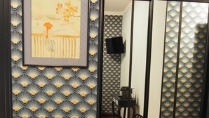 Una habitación con suelo de madera, pared con un diseño negro y dorado, una obra de arte enmarcada, una mesita con un jarrón y una puerta corredera con un dibujo de abanicos.