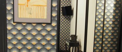 A room with a wooden floor, a black and gold patterned wall, a framed artwork, a small table with a vase, and a sliding door with a fan pattern.