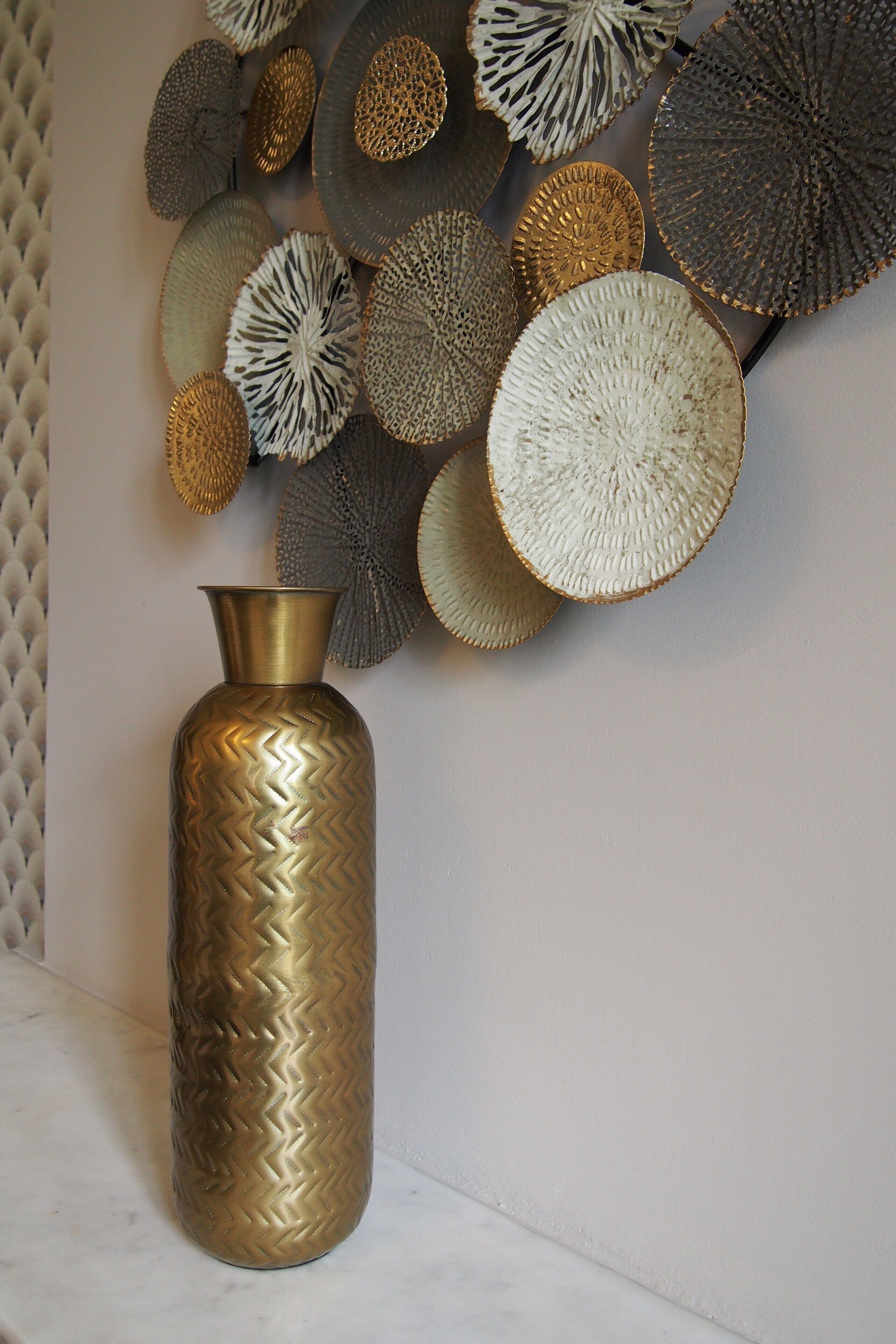 Una pared decorativa con diferentes placas texturizadas y un jarrón de latón.