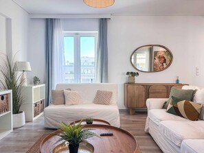 A modern living room with a sofa, a coffee table, a wooden cabinet, and a large window with curtains. - Boho Flat in Glyfada Center (Glyfada)