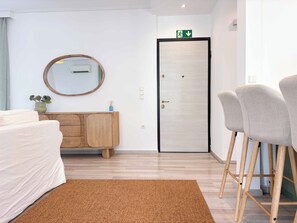 A modern hotel room with a wooden cabinet, a round mirror, a beige sofa, a brown rug, and a door with an emergency exit sign. - Boho Flat in Glyfada Center (Glyfada)
