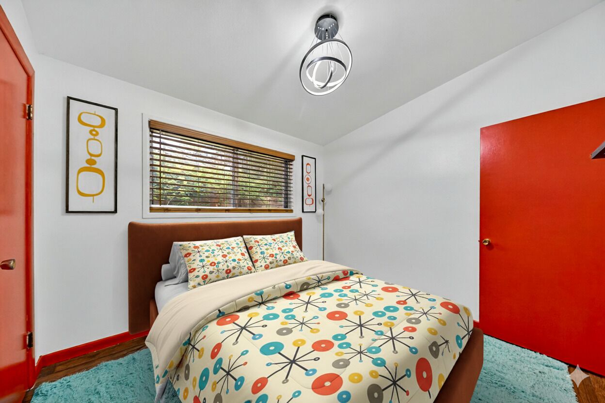 A bedroom with a bed, a window with blinds, a red door, and a ceiling light fixture.