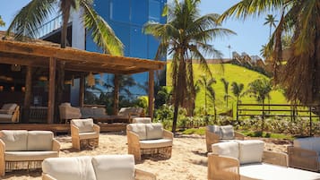A beachside lounge area with wicker furniture and a view of a blue building and palm trees.