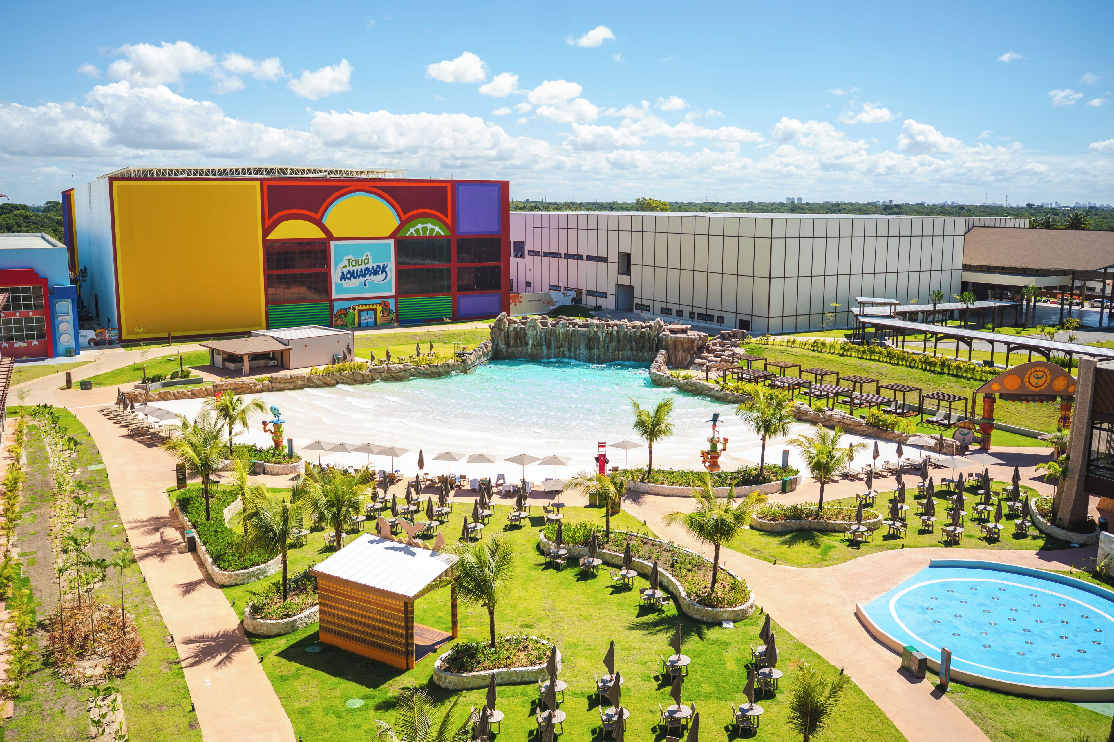Resort com uma piscina grande, áreas de estar ao ar livre e um edifício colorido com design de sol e ondas.