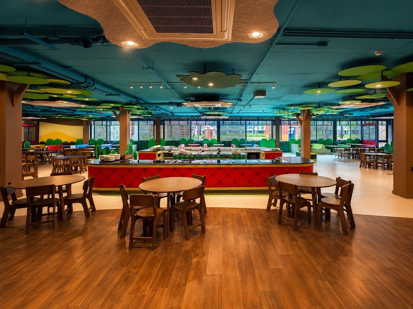 A spacious indoor dining area with wooden tables and chairs, a central counter with a red accent, and large windows providing natural light.