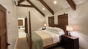 Une chambre avec un plafond en pente, des poutres en bois, un lit à baldaquin, une table de chevet avec une lampe, et une salle de bain à l’arrière-plan.