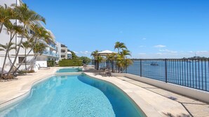 A swimming pool with a curved design, surrounded by palm trees and a view of the sea. - Commodore 6 Noosa Heads (Noosa Heads)