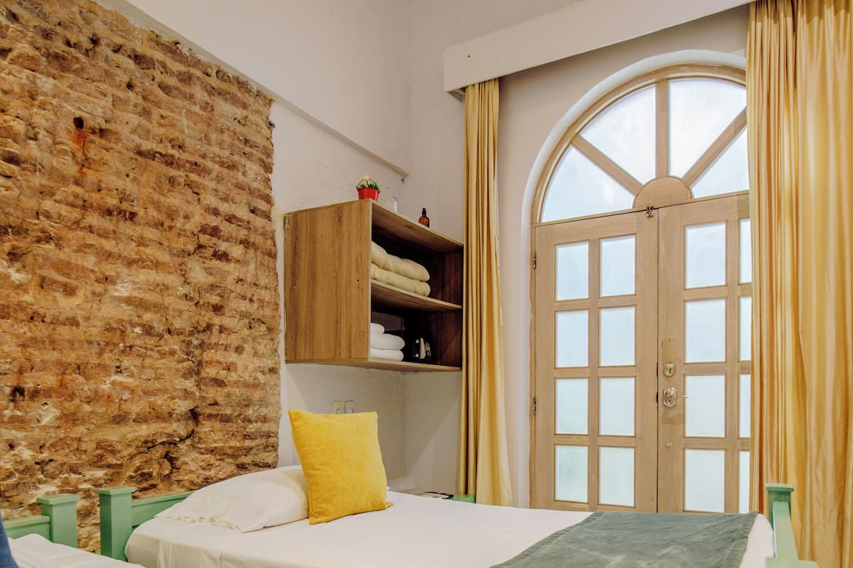 Une chambre avec deux lits, un mur en briques, une armoire en bois et une grande fenêtre en arc surmontée de rideaux.