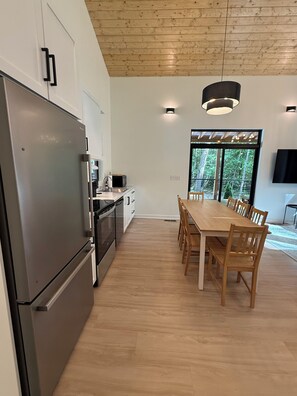 A modern kitchen with a large refrigerator, stove, and microwave. There is a dining area with a wooden table and chairs. - Creekside Nook in Sewanee (Sewanee)
