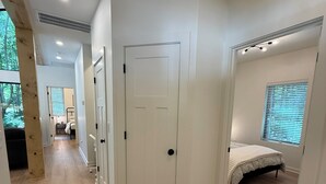 Un couloir doté d’un plancher en bois, de murs blancs et d’une porte donnant sur une chambre à coucher, où se trouvent un lit et des stores aux fenêtres.