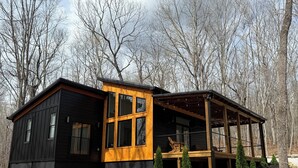 Une cabane à l’extérieur noir et dotée d’une terrasse en bois, entourée d’arbres.
