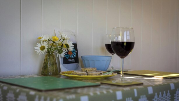 A table with a glass of red wine, a vase with flowers, a brochure, and a plate with food items.