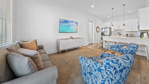 Un salon moderne avec un canapé, un fauteuil à motifs et une table basse. On y trouve également un coin cuisine avec un bar et des meubles blancs.
