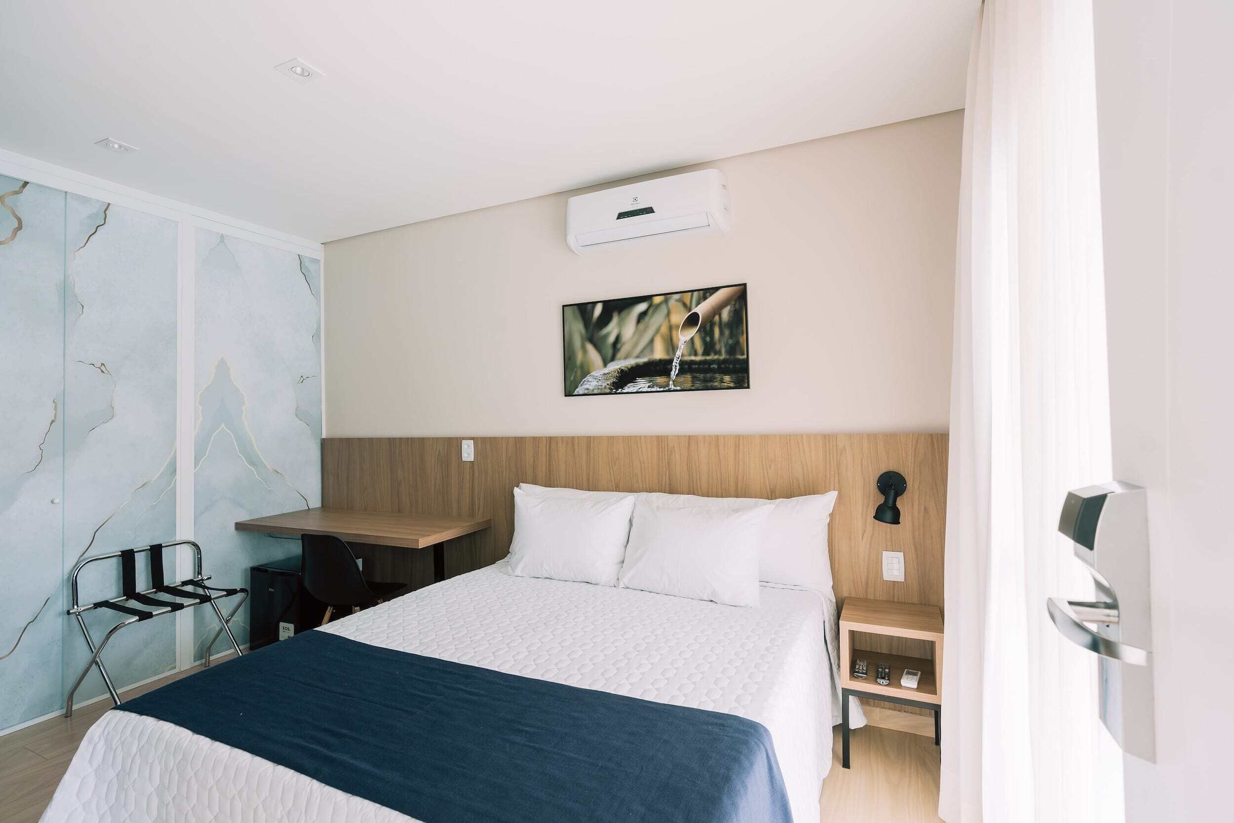 A hotel room with a bed, a desk, a chair, a small table, and a picture on the wall.