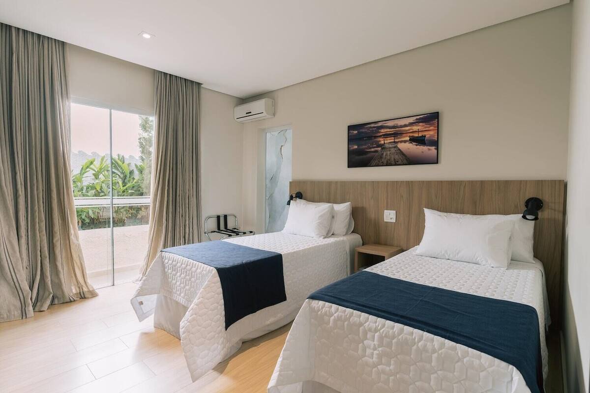 A hotel room with two beds, a large window with curtains, an air conditioning unit, and a painting on the wall.