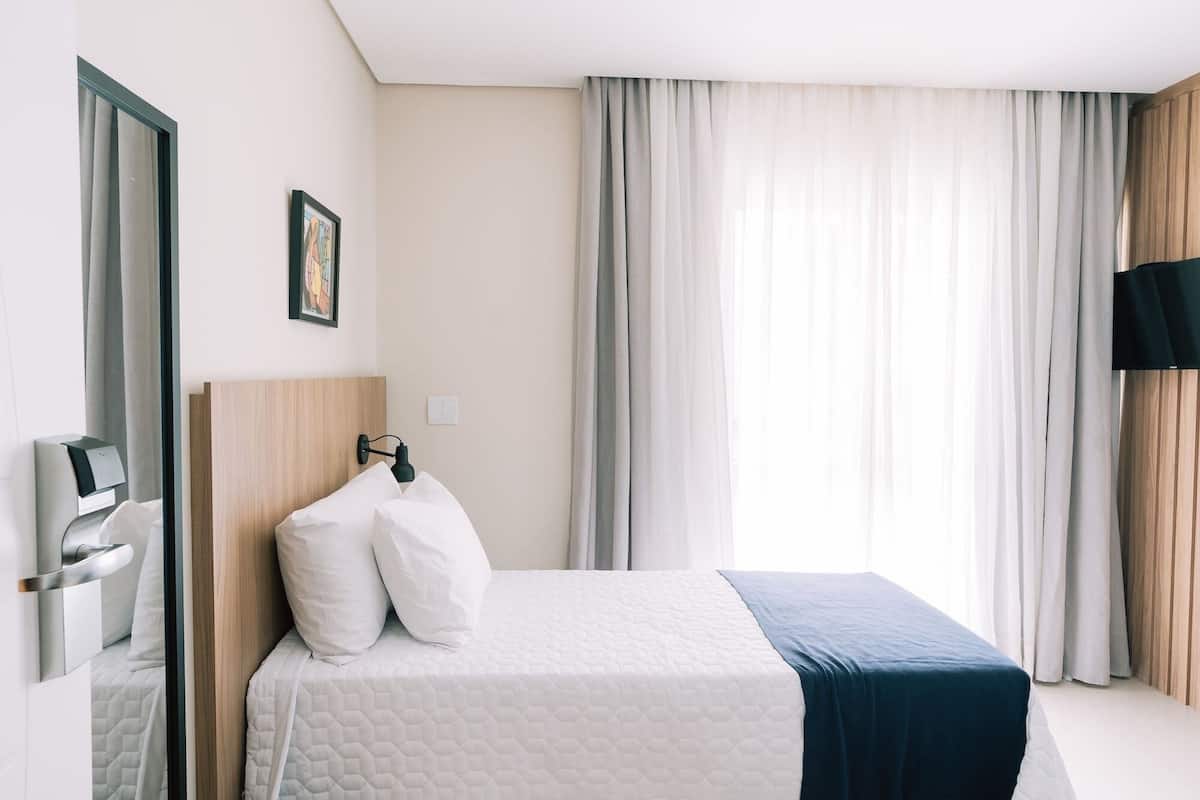A hotel room with a bed, a headboard, a wall-mounted TV, and a window with curtains.