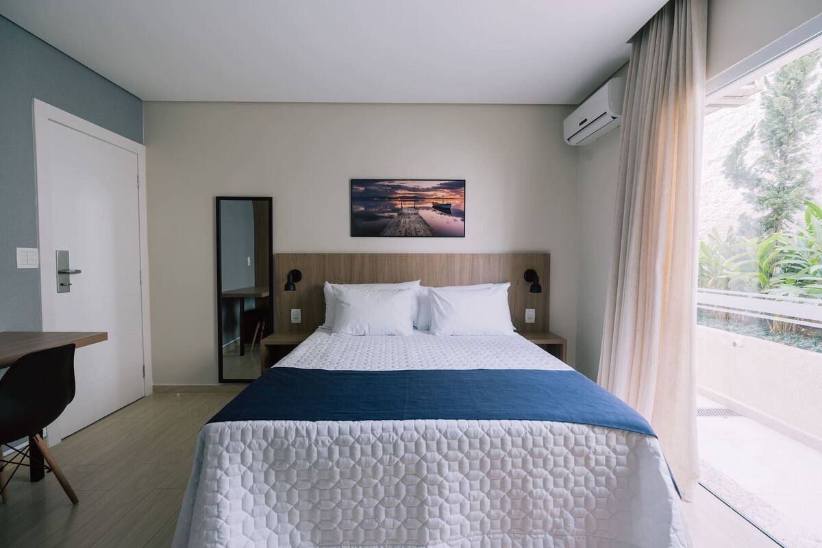A hotel room with a bed, a desk, a chair, a painting on the wall, and a window with curtains.