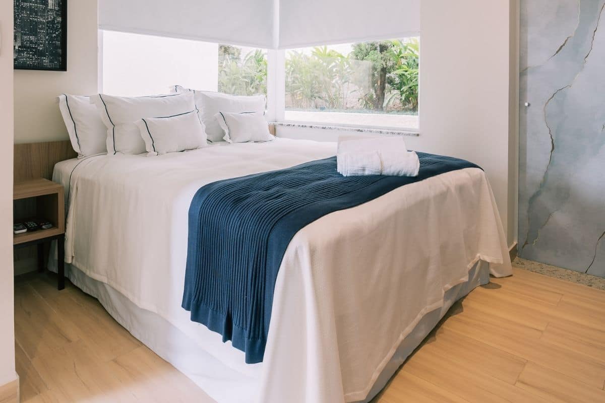A neatly made bed with white and blue bedding, a wooden nightstand, and a window with a view of greenery.