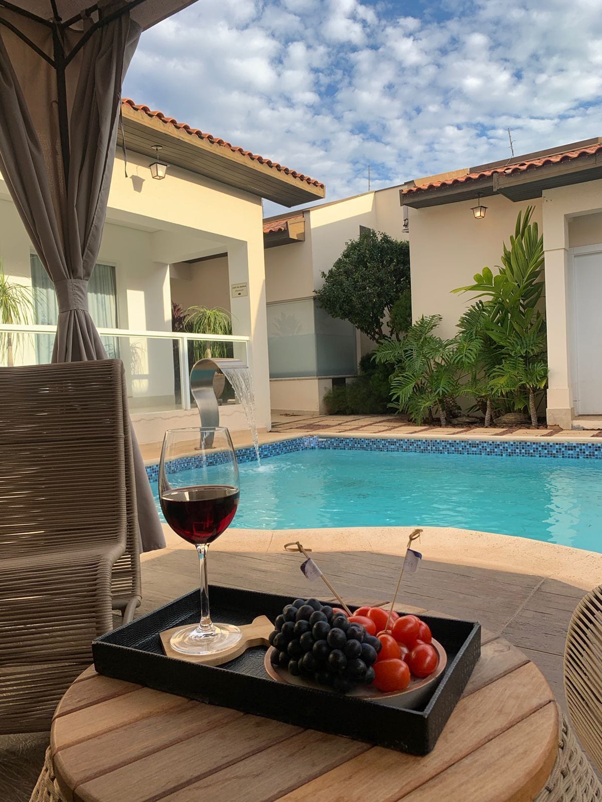 Escena junto a la piscina con una copa de vino tinto, una bandeja con uvas y tomates, y una silla de mimbre.