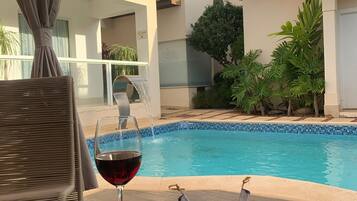 Escena junto a la piscina con una copa de vino tinto, una bandeja con uvas y tomates, y una silla de mimbre.
