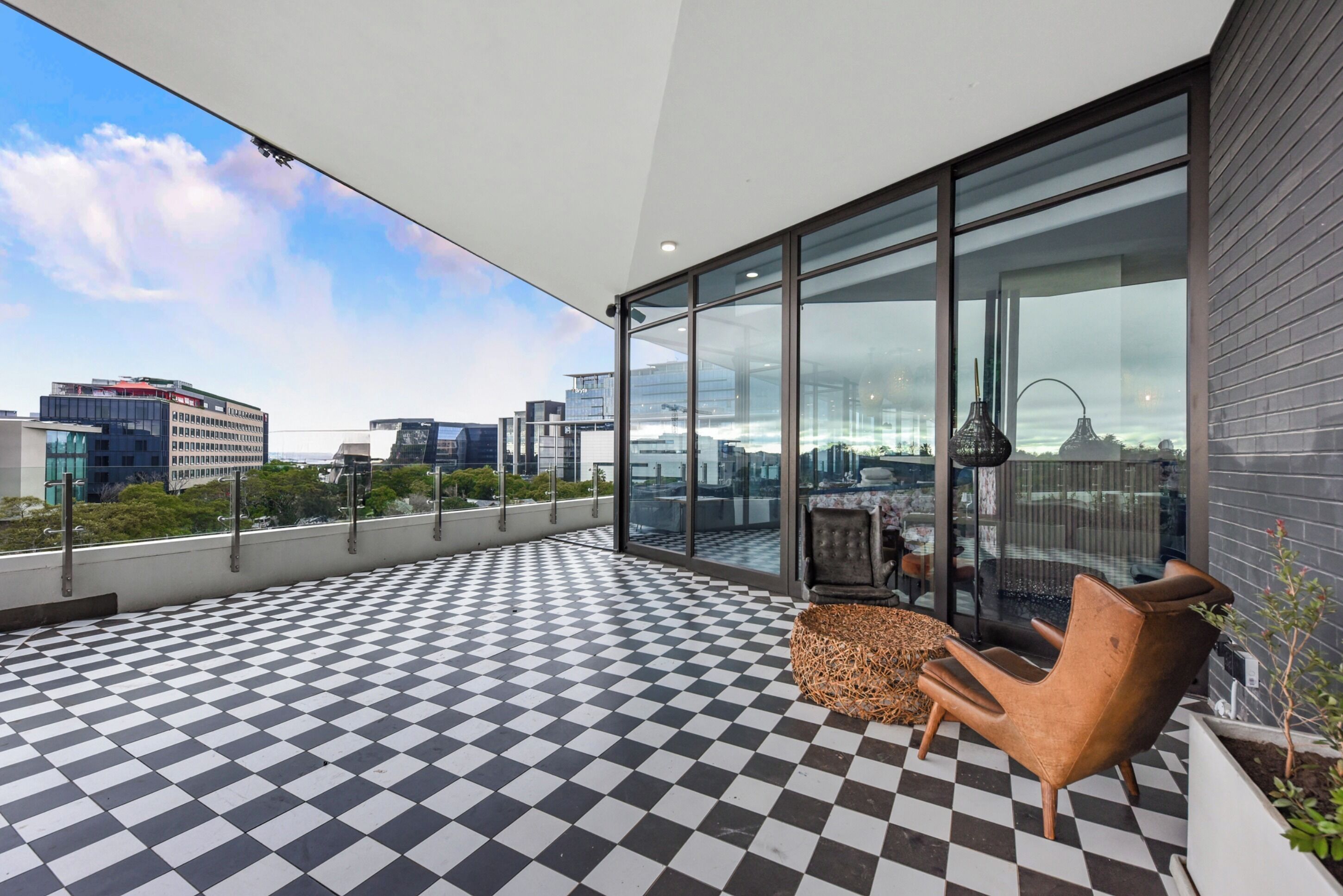 A rooftop terrace with a checkered floor, a seating area with a chair and a table, and a glass wall offering a city view.