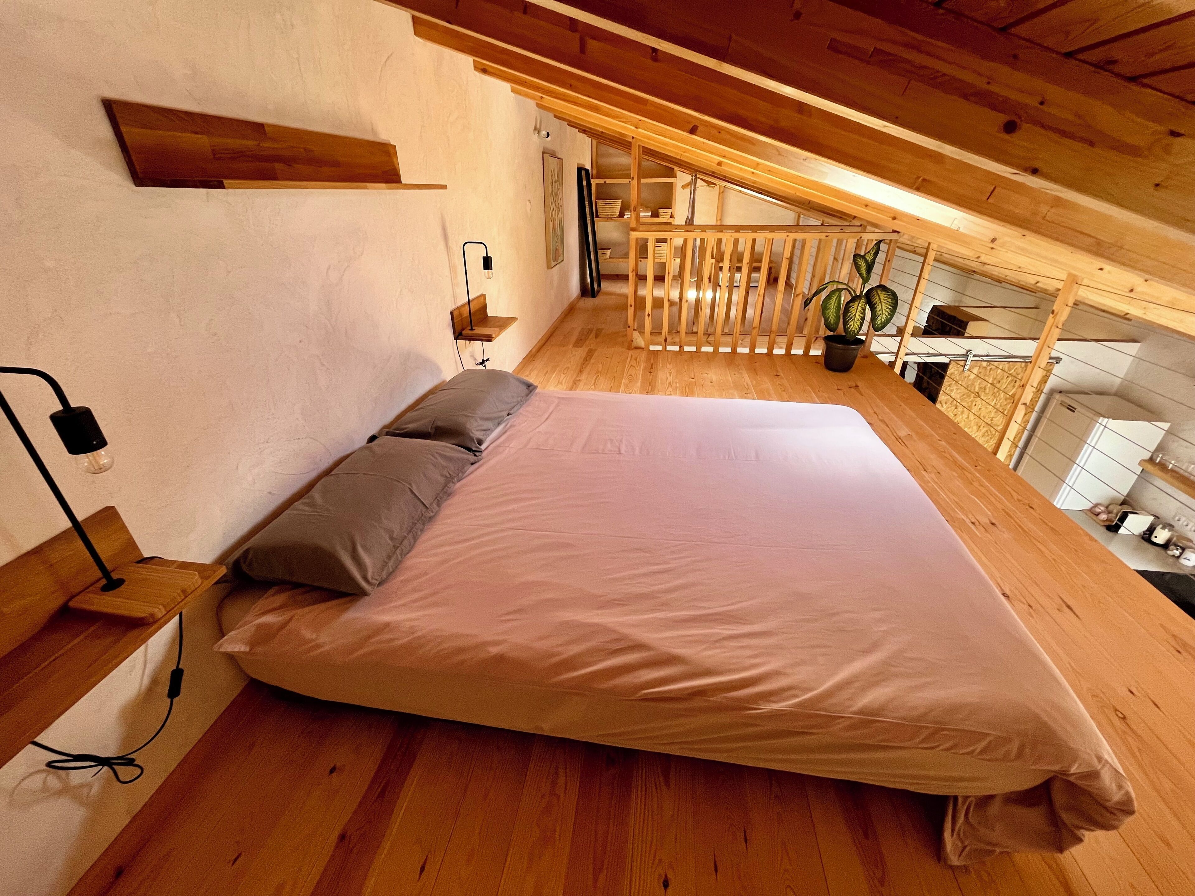 A bedroom with a large bed, wooden floor, a sloped ceiling, and a small balcony with plants.
