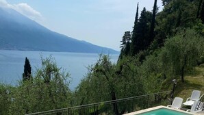Une piscine rectangulaire à l’eau d’un bleu limpide, entourée d’une terrasse carrelée. On y trouve des chaises longues blanches et une petite table. La piscine est située devant un décor composé d’arbres et d’une chaîne de montagnes lointaine, et on aperçoit une étendue d’eau à l’arrière-plan.