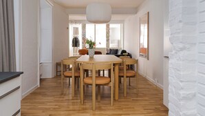 A dining area with a round wooden table and six chairs, a hanging light fixture, and a view into another room with a sofa and a window. - Kings48 by Daniel&Jacob's (Copenhagen)