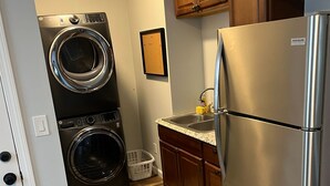A compact kitchen with a stainless steel refrigerator, a double washer-dryer unit, wooden cabinets, and a striped rug.